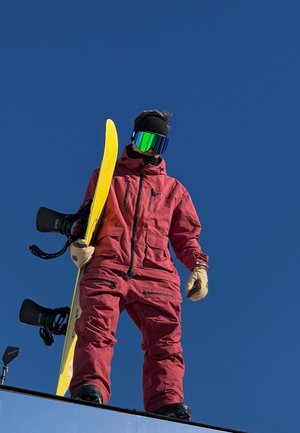 Esquí con un traje aislante rojo, tabla de snowboard amarilla con una ligera curvatura, guantes negros, gafas de espejo coloridas, de pie sobre una superficie nevada bajo un cielo azul claro.