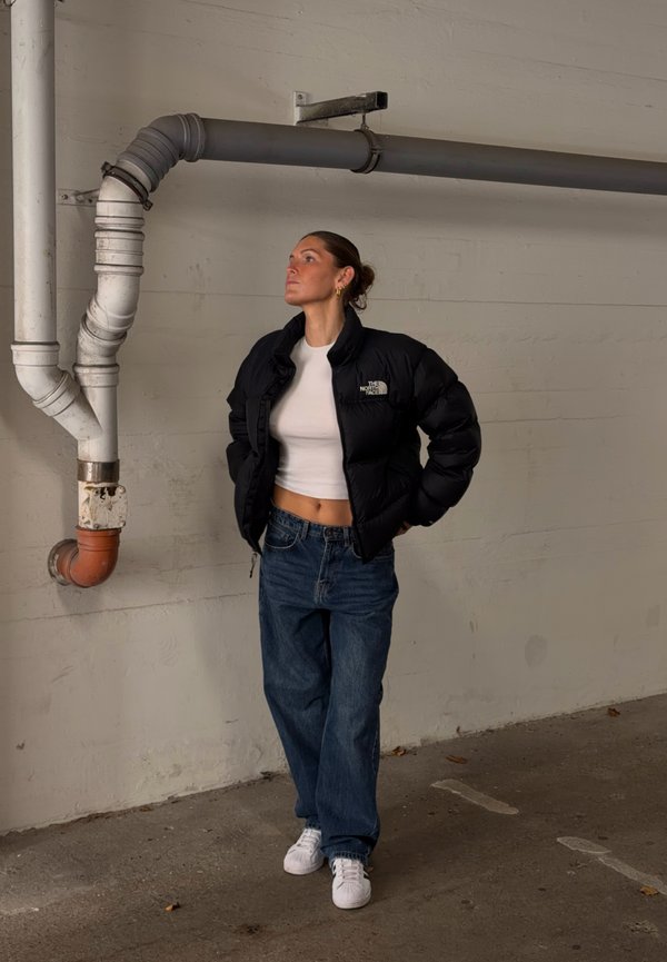 Una mujer luce una chaqueta acolchada negra de The North Face, un top corto blanco, unos vaqueros holgados y unas zapatillas blancas en una sala de estilo industrial.
