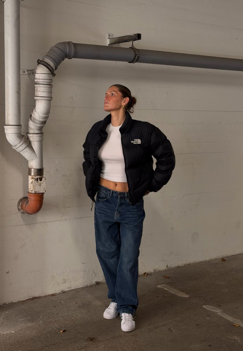 Une femme porte une doudoune noire The North Face, un crop top blanc, un jean ample et des baskets blanches dans une pièce au style industriel.