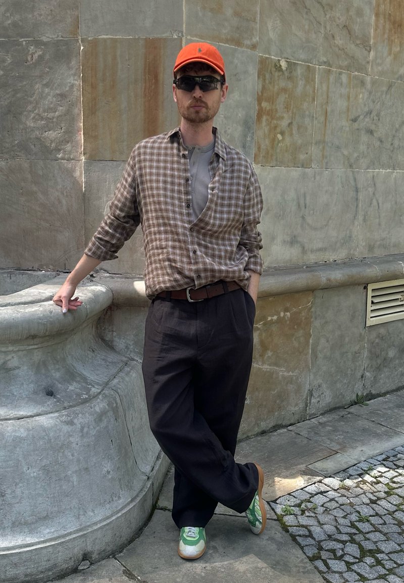 Homme portant une casquette orange, des lunettes de soleil sombres, une chemise à carreaux et un pantalon foncé, appuyé contre un mur en pierre à l’extérieur, une jambe croisée.