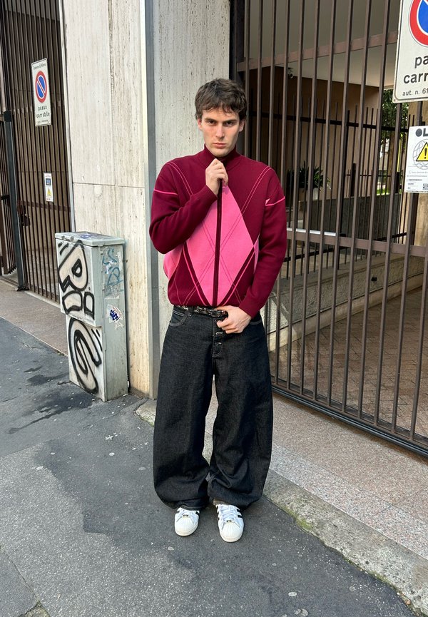 Jeune homme debout sur le trottoir, portant un pantalon noir trop grand, des baskets blanches et un pull à losanges bordeaux et rose, devant des grilles métalliques et des panneaux de rue urbains.