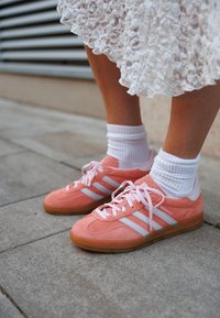Ροζ σουέτ αθλητικά παπούτσια με λευκές ρίγες και κορδόνια, με υφή στο πάνω μέρος και σόλα από καουτσούκ, φορεμένα με λευκές ριμπ κάλτσες.
