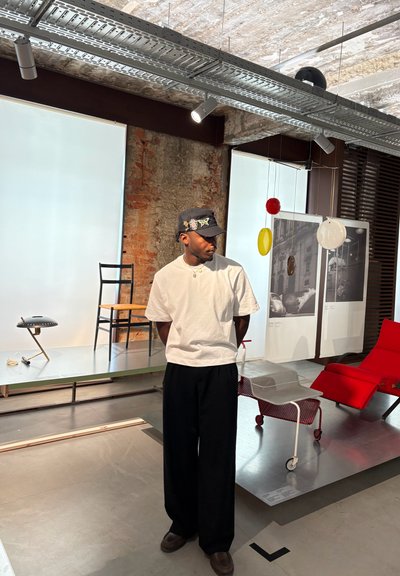 Una persona está de pie en un espacio de exhibición vistiendo una camisa blanca y pantalones negros, con una silla, una lámpara y un reclinable rojo al fondo.