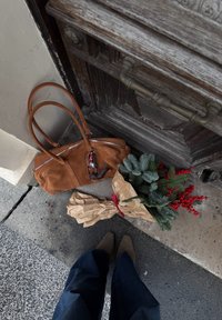 Sac à main en daim marron avec deux poignées et une fermeture éclair argentée, à côté d'un bouquet enveloppé de feuillage persistant et de baies rouges sur une surface texturée.