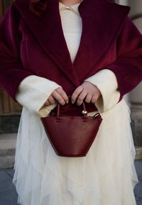 Bordó bőrből készült kézitáska háromszög alakú, kis fogantyúkkal és arany díszítéssel. Fehér réteges ruhával és bordó kabáttal viselik.