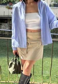 Ανοιχτό μπλε πουκάμισο με κουμπιά, λευκή τοπ χωρίς τιράντες, χακί μίνι φούστα με τσέπη στο πλάι, πράσινη τσάντα, μαύρες μπότες μέχρι το γόνατο, εξωτερικό σκηνικό δίπλα στο νερό.