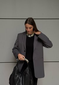 Szürke, szabott blézer fekete pulóver fölött, gyöngy nyaklánccal párosítva, és egy texturált fekete kézitáskát tartva, ezüst fogantyúval.