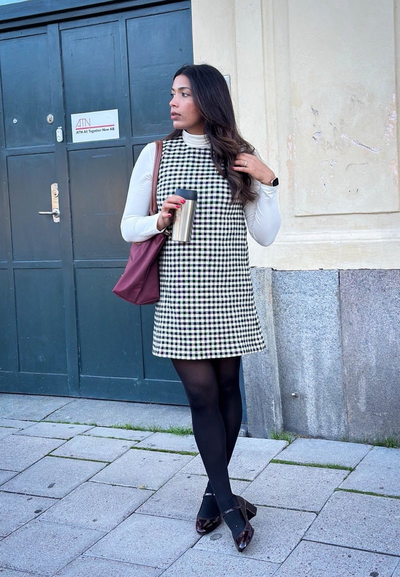 Checkered dress in black and white, sleeveless with a mock neck, paired with black tights and brown shoes. Holding a metal travel mug.