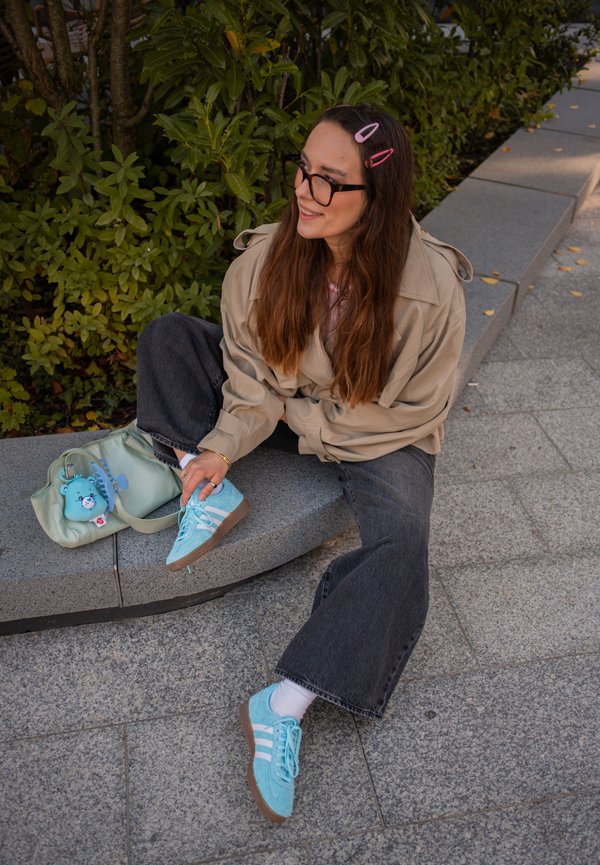 Jeune femme aux longs cheveux bruns et lunettes assise sur un banc en pierre à l'extérieur, portant une veste beige, un pantalon noir large et des baskets bleu vif.
