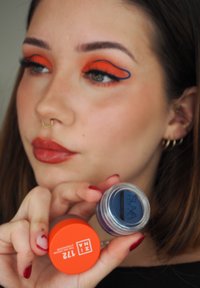 Contenitore arancione etichettato "INNA 712" e vaso cosmetico blu etichettato "SUVA" tenuti da una persona con trucco occhi arancione e blu audace.
