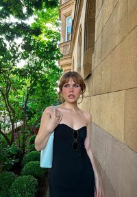 Μαύρο strapless φόρεμα με εφαρμοστή σιλουέτα, συνδυασμένο με μία ανοιχτό μπλε τσάντα. Τα αξεσουάρ περιλαμβάνουν στρογγυλά γυαλιά ηλίου και χρυσά σκουλαρίκια.