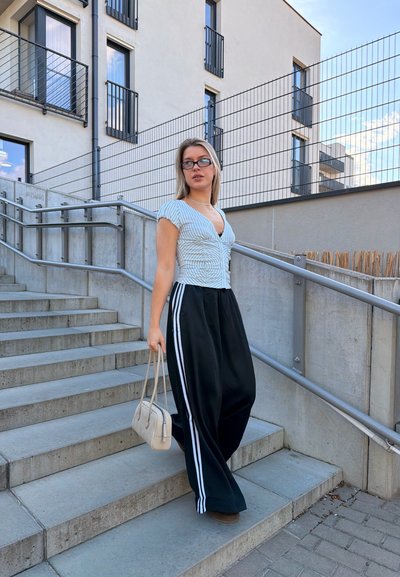 Joven mujer con gafas, top de rayas, pantalones negros anchos con rayas blancas, sujetando un bolso beige, bajando unas escaleras exteriores de hormigón.