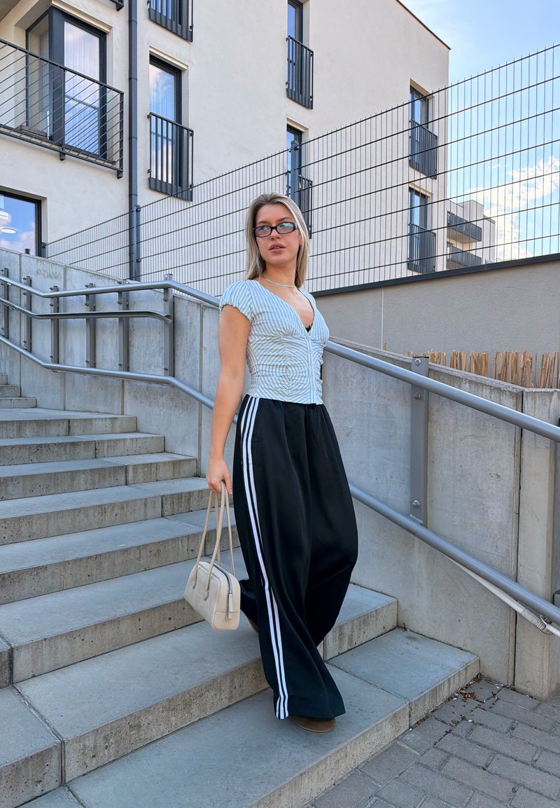 Jeune femme portant des lunettes, haut rayé, pantalon large noir avec des rayures blanches, tenant un sac à main beige, marchant dans des escaliers extérieurs en béton.