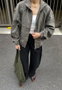 Nadměrná olivově zelená bunda se zipem vpředu, texturovaná látka, elastické manžety, bílý tílko, černé široké kalhoty a hnědé mokasíny.