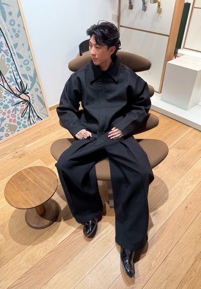 Chaqueta y pantalones negros oversize de tela texturizada, usados con zapatos negros, sentado en un taburete redondo marrón junto a un fondo estampado.