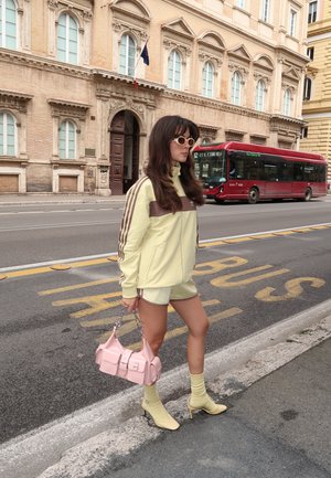 Jasnożółta kurtka sportowa z brązowymi akcentami, dopasowane szorty, torebka w pastelowym różu i żółte botki na obcasie. Scena uliczna z autobusem.