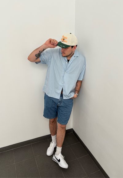 Camisa de rayas azul claro de manga corta, pantalones cortos de mezclilla, zapatillas blancas con detalles negros y una gorra crema con visera verde y gráficos.
