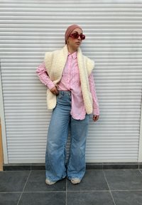 Colete creme fofo sobre uma camisa rosa às riscas com botões, combinado com jeans de perna larga azul-claro e sapatos bege. Acessórios incluem grandes óculos de sol vermelhos.