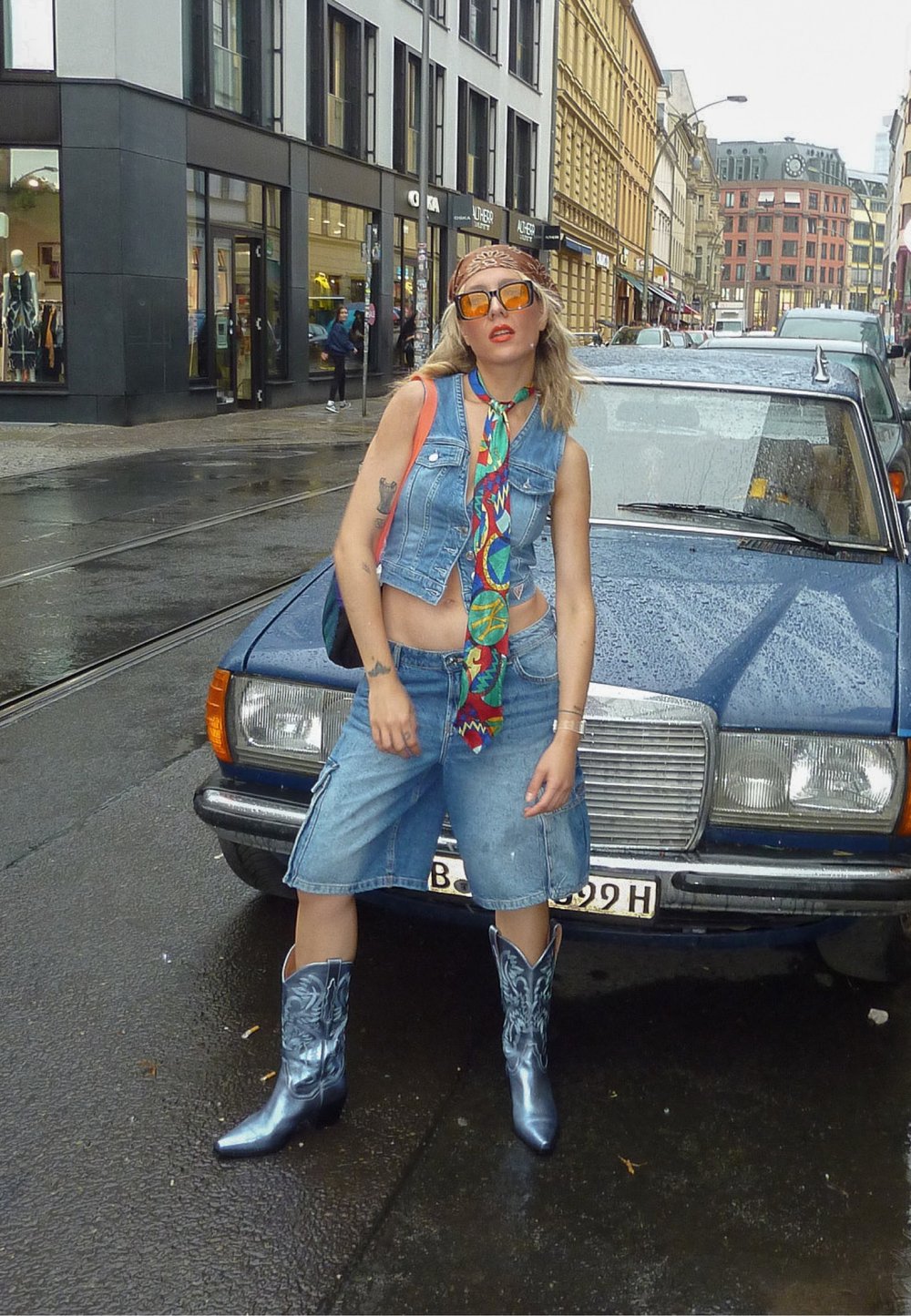 Denim brezrokavnik in kratke hlače, večbarvni vzorčasti šal, sijoče modre kavbojske čevlje, urbano ulico s modrim avtomobilom in mokrim pločnikom.