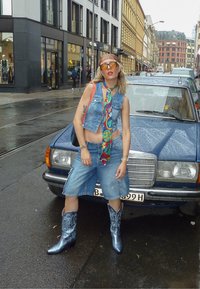 Denim brezrokavnik in kratke hlače, večbarvni vzorčasti šal, sijoče modre kavbojske čevlje, urbano ulico s modrim avtomobilom in mokrim pločnikom.