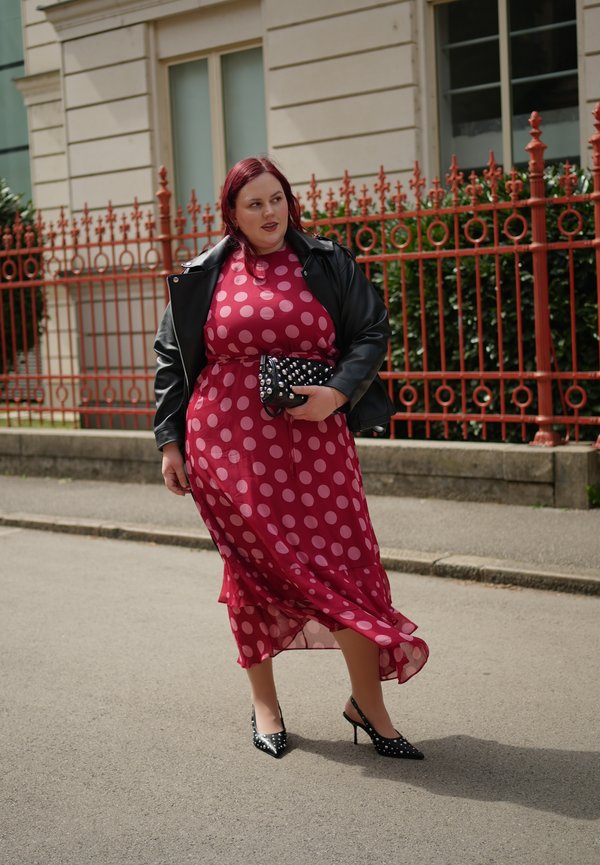 Femme marchant à l'extérieur portant une robe rouge à pois, une veste en cuir noire, tenant une pochette cloutée et des talons noirs cloutés.