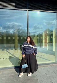 Mujer con cabello largo y rizado que se encuentra afuera de un edificio de cristal, vestida con un suéter a rayas, falda ancha negra y zapatos blancos, sosteniendo un bolso azul claro.