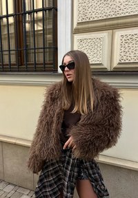 Femme portant une grande veste en fausse fourrure marron, une jupe à carreaux et des lunettes de soleil noires, debout contre un mur orné d'un bâtiment beige avec des fenêtres grillagées.