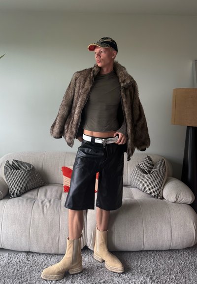 Chaqueta de piel sintética marrón, camiseta ajustada verde oliva, pantalones cortos de cuero negros, botas beige hasta el tobillo con suelas de goma y una gorra negra y roja.