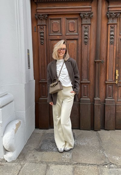 Abrigo de lana marrón sobre una camisa blanca, pantalones de pernera ancha color crema y zapatos grises. Pequeño bolso gris con un cierre dorado. Puerta de madera en el fondo.