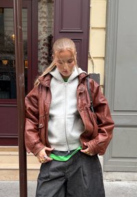 Brun skinnjakke i stor størrelse over en grå hettejakke med glidelås, grønn skjorte og løse grå bukser. Merkbare ringer på hendene. Urban bakgrunn synlig.