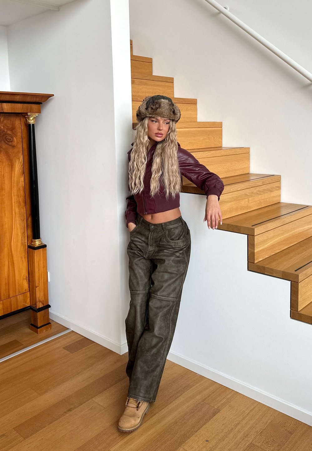 Brun läderkappa i cropped-modell med matchande pälshatt, olivgröna vida byxor och beige stövlar; mot en bakgrund av trätrappor och detaljerad vägg.