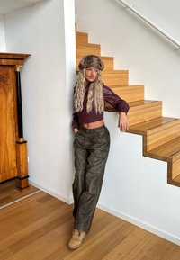 Brun läderkappa i cropped-modell med matchande pälshatt, olivgröna vida byxor och beige stövlar; mot en bakgrund av trätrappor och detaljerad vägg.