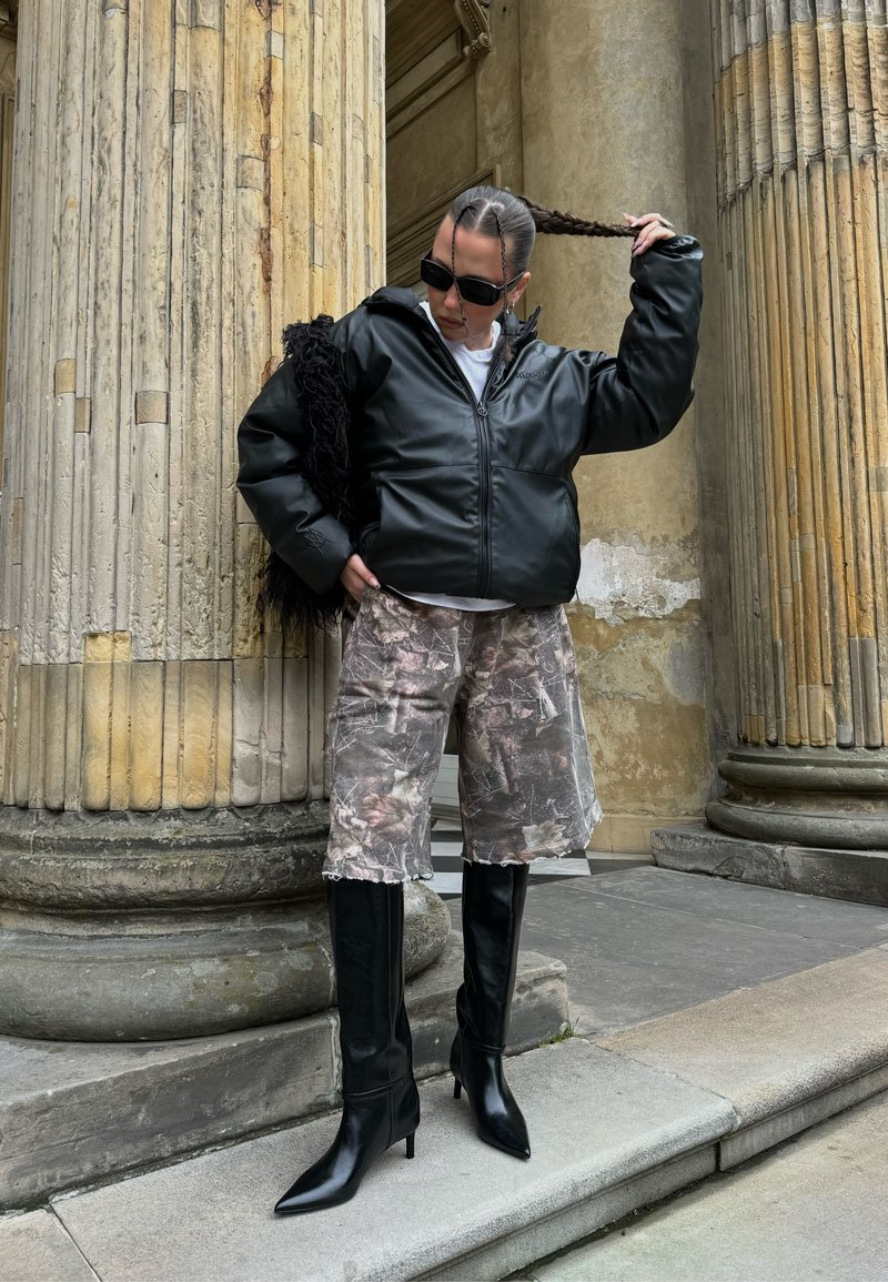 Giacca nera in pelle con zip, pantaloncini con stampa mimetica e stivali neri alti sopra il ginocchio con punte appuntite. Sfondo testurizzato di colonne in pietra.