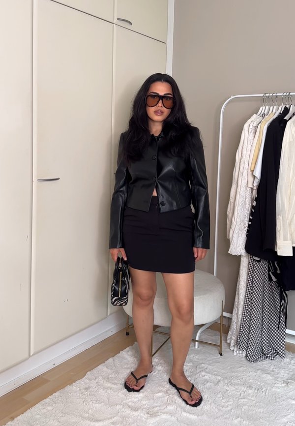 Mujer con cabello largo y oscuro, que lleva gafas de sol grandes, chaqueta de cuero negra, falda negra, sosteniendo un bolso pequeño en un dormitorio con perchero para ropa.