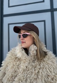 Femme aux cheveux blonds portant une casquette sombre, des lunettes de soleil roses et un manteau beige duveteux, se tenant devant un mur à panneaux bleu-gris.