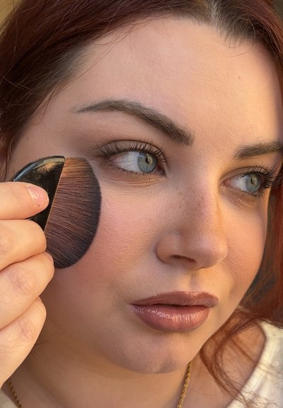 Brocha de maquillaje con mango negro y cerdas suaves usada en la mejilla. La persona tiene piel clara, pestañas marcadas y color de labios neutro.