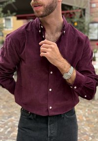 Bordeauxkleurig overhemd met knopen en een ribfluweelstructuur, voorzien van witte knopen en opgerolde manchetten, gecombineerd met zwarte spijkerbroek en een zilveren horloge.