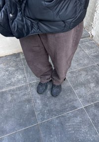 Μαύρο επενδυμένο μπουφάν, καφέ παντελόνια σε άνετη γραμμή και μαύρα αθλητικά παπούτσια με υφή, που στέκονται σε γκρι πλακάκια δαπέδου.