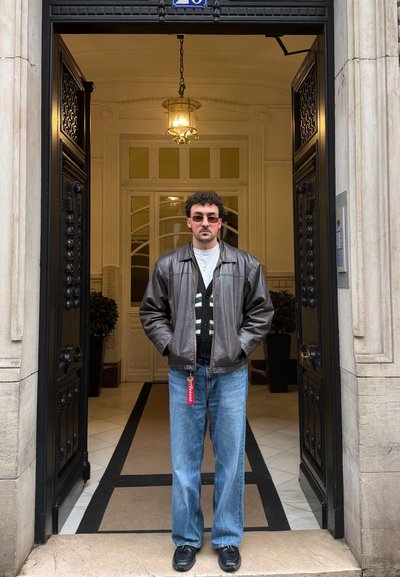 Hombre con gafas de sol, chaqueta de cuero negra, suéter con estampado, vaqueros azules y zapatos negros, de pie en el marco abierto de la puerta de un edificio.