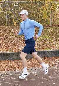 Sportiskais tērps ietver gaiši zilu garroku kreklu, tumši zilas šortus, baltas zeķes un skriešanas apavus ar melnām svītrām. Fona atrodas rudens lapas.