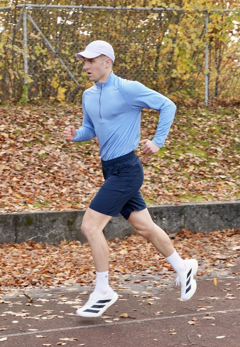 Completo sportivo con maglia a maniche lunghe azzurro chiaro, pantaloncini blu navy, calzini bianchi e scarpe da corsa con strisce nere. Foglie autunnali sullo sfondo.