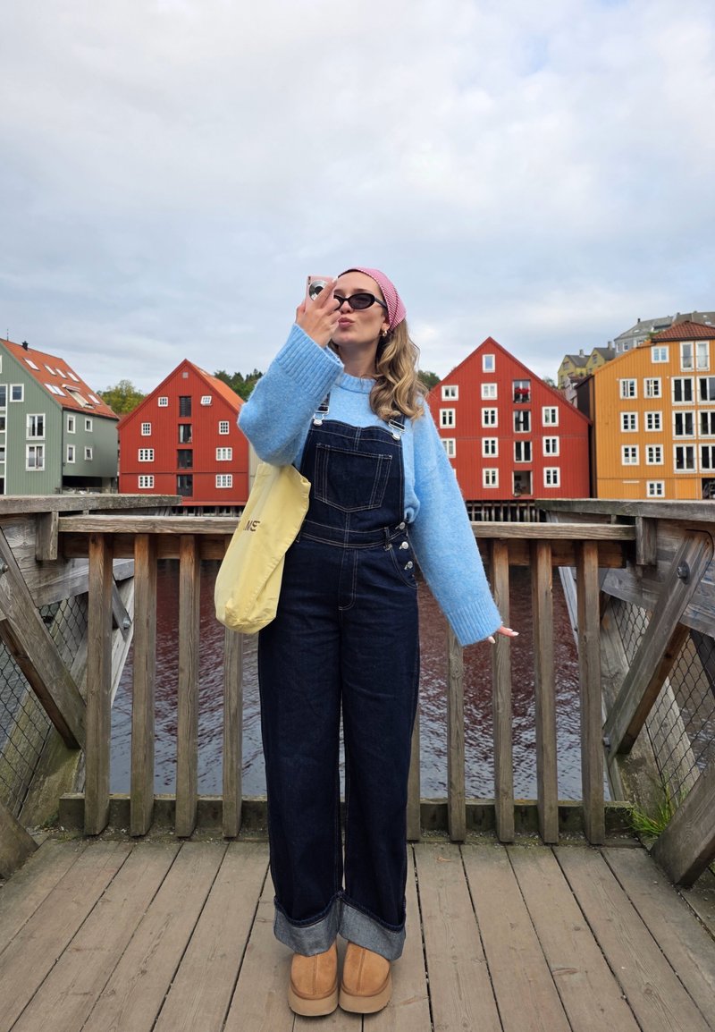 Un pull bleu clair et moelleux associé à des salopettes en denim bleu foncé. Des chaussures à plateforme beige et un sac fourre-tout jaune. En arrière-plan, des bâtiments colorés.