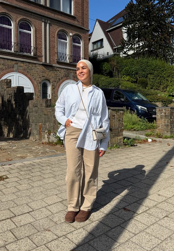 Woman wearing a white hijab, striped shirt, beige pants, and brown shoes stands smiling on a sunlit paved street in a residential area.