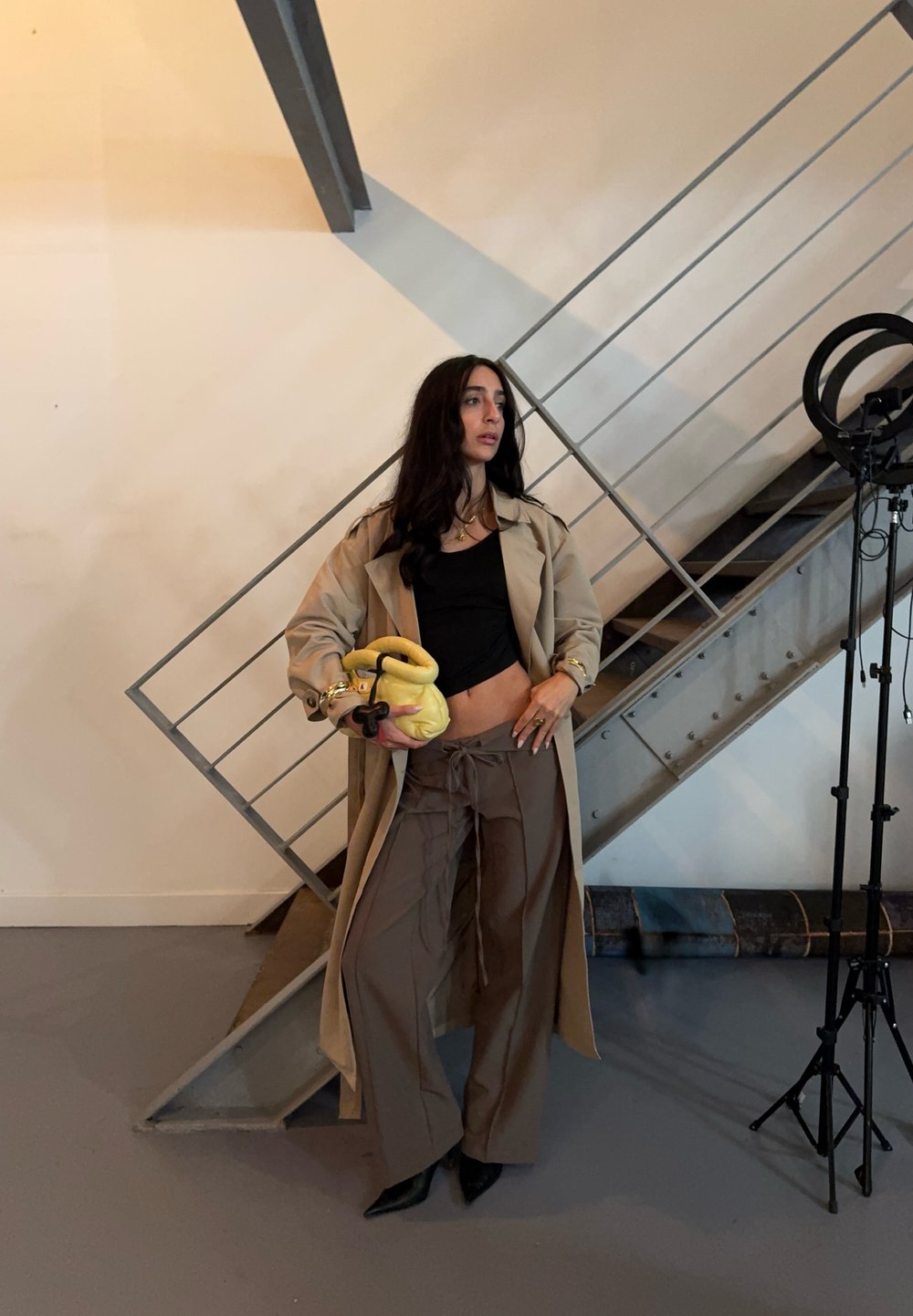 Bež trench coat, must crop top, pruunid laiad püksid, kollane käekott, mustad kontsad. Seisab metalltrepi ja valgustusvarustuse lähedal.