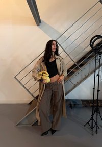 Bež trench coat, must crop top, pruunid laiad püksid, kollane käekott, mustad kontsad. Seisab metalltrepi ja valgustusvarustuse lähedal.