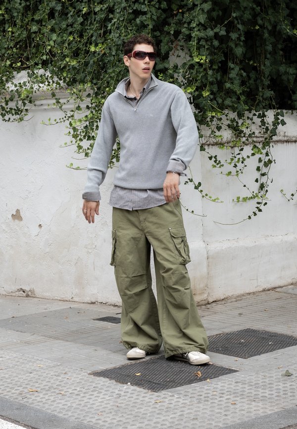 Joven con gafas de sol, suéter gris y pantalones cargo verdes sueltos parado en una acera con una pared blanca y hiedra al fondo.