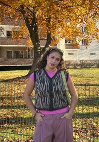 Veste tricotée rayée noire et brune portée sur une chemise rose à manches courtes, associée à un pantalon large violet clair et un sac à épaule vert.