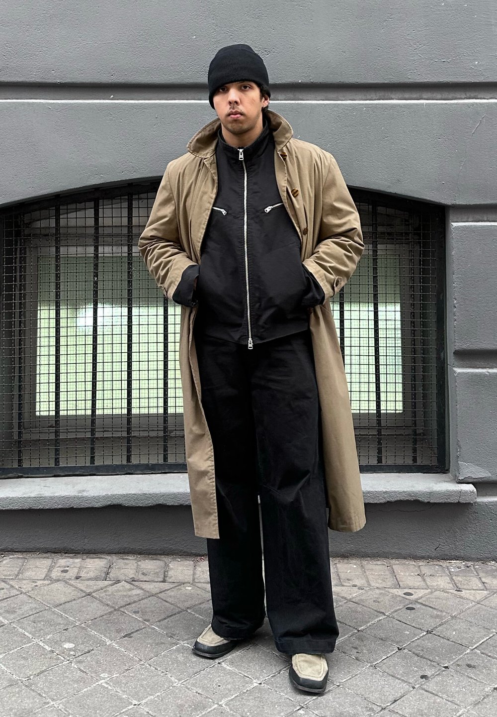 Νέος άνδρας που φοράει μπεζ καμπαρντίνα, μαύρο μπουφάν, μαύρο παντελόνι, σκουφάκι και ανοιχτόχρωμα παπούτσια, στέκεται στο πεζοδρόμιο της πόλης μπροστά σε γκρι τοίχο.