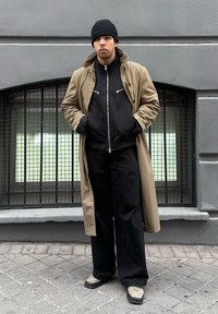 Νέος άνδρας που φοράει μπεζ καμπαρντίνα, μαύρο μπουφάν, μαύρο παντελόνι, σκουφάκι και ανοιχτόχρωμα παπούτσια, στέκεται στο πεζοδρόμιο της πόλης μπροστά σε γκρι τοίχο.