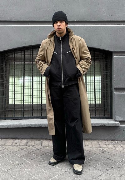 Joven con abrigo beige, chaqueta negra, pantalones negros, gorro y zapatos de color claro, de pie en la acera de la ciudad junto a una pared gris.